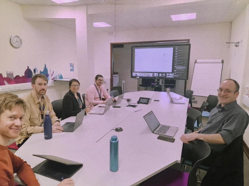 Team pic Image of 5 team members sitting around a meeting room table, with laptops and smiling at the camera